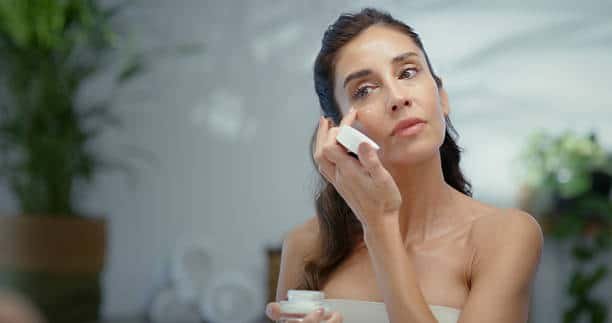 gettyimages-2211749265-612×612 Skincare, face cream and woman by mirror in bathroom for natural, moisturizing or hydration routine. Happy, reflection and female person with facial spf, sunscreen or lotion for dermatology in home.